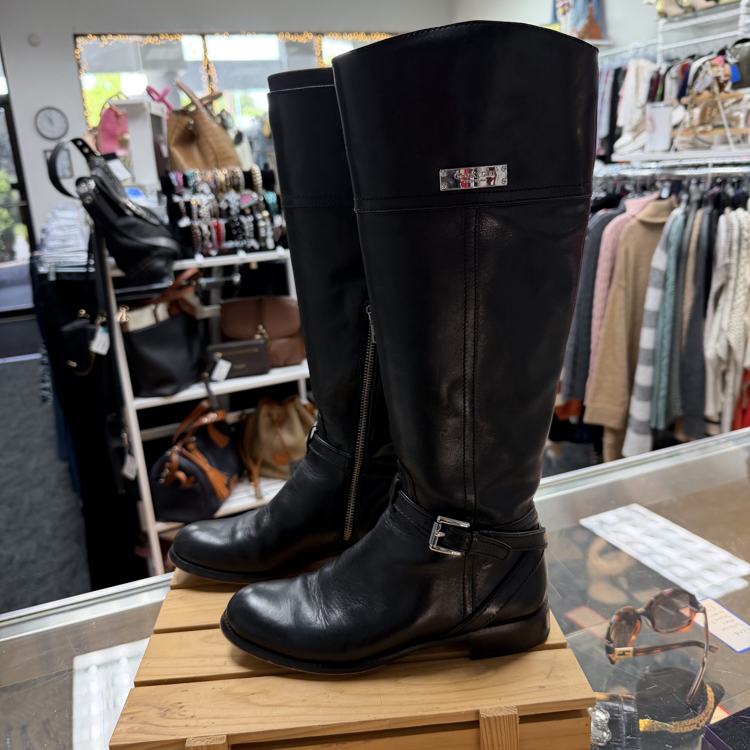 Authentic Coach black leather riding boots with buckle detail and silver-tone Coach hardware displayed in a women's consignment boutique.